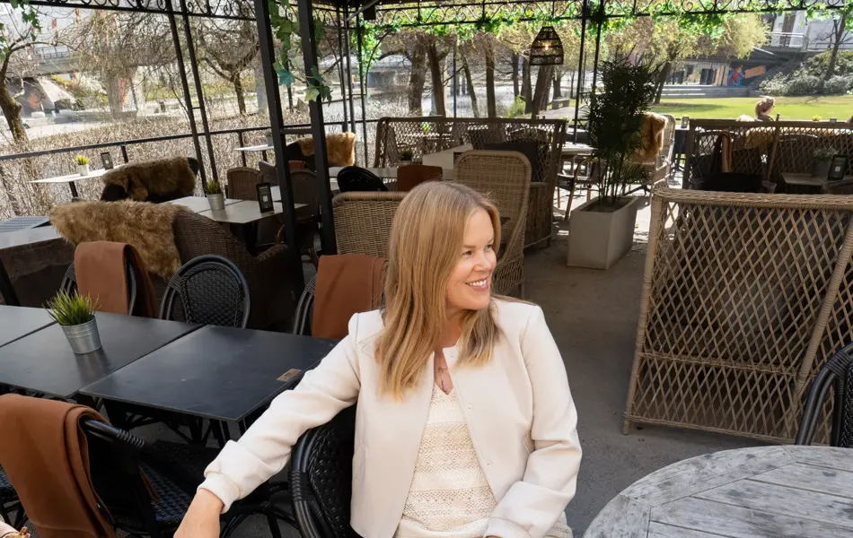 Dame sitter på cafe med beige kort jakke, beige strikket kjole og brunmønstret sandaler