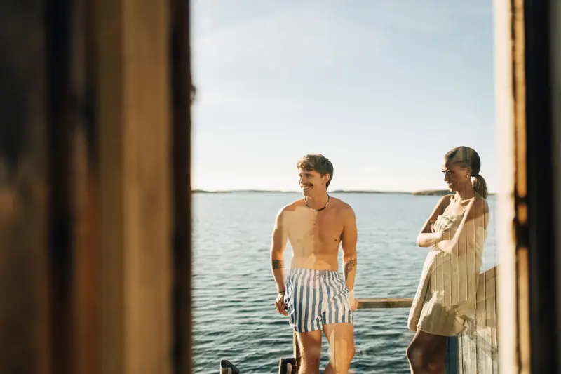 Mann med hvit- og blåstripete shorts står utenfor badstue med havet i kveldssol i bakgrunnen