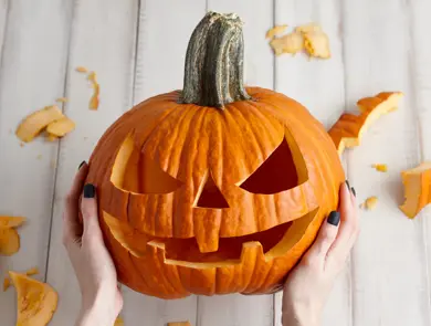 Woman carving big orange pumpkin into jack-o-lantern for Halloween holiday decoration on white wooden planks, close up view