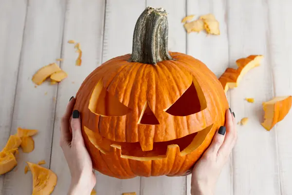 Woman carving big orange pumpkin into jack-o-lantern for Halloween holiday decoration on white wooden planks, close up view