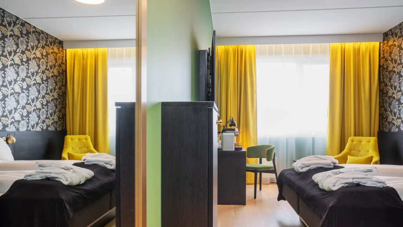 Stort speil, grønn vegg, skrivebord med stol, gule gardiner, gul stol og morgenkåper på sengen i superior rom på Thon Hotel Kirkenes