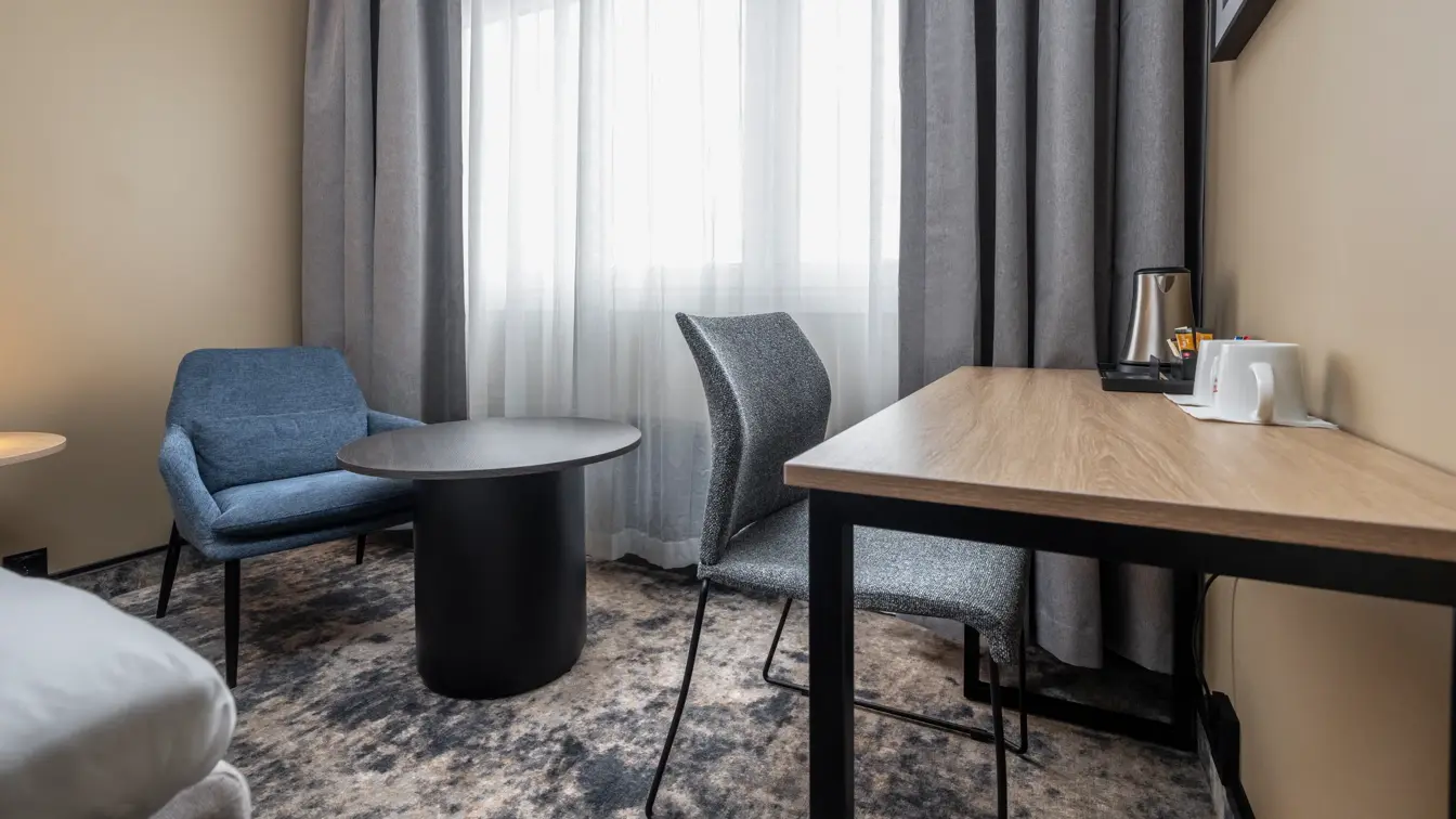 Dobbeltrom med lenestol og tilhørende bord, skrivebord med stol, lange gardiner og stort vindu.