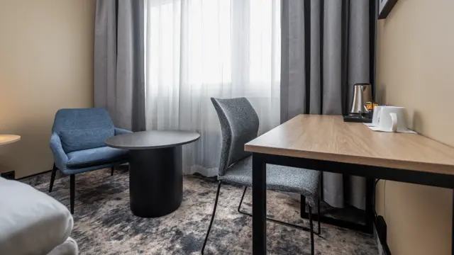 Dobbeltrom med lenestol og tilhørende bord, skrivebord med stol, lange gardiner og stort vindu.