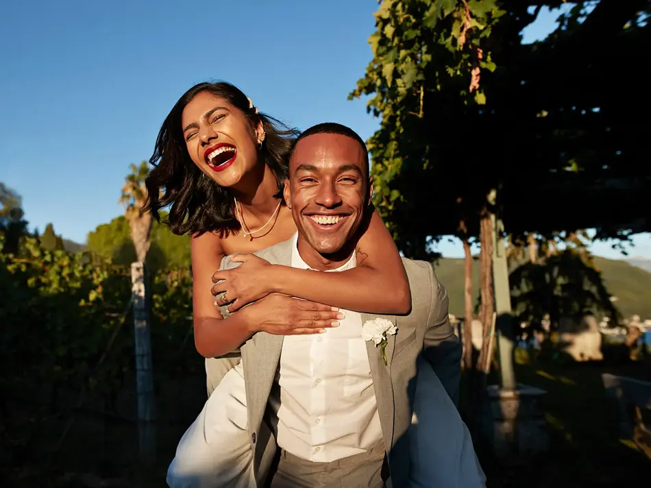Smilende dame sitter på ryggen til mann i hvit skjorte og beige dressjakke