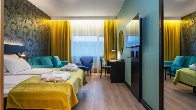 Parkettgulv, turkis sofa, to stoler, salongbord, skrivebord, dobbeltseng, vindu fra gulv til tak, gule gardiner, tapetvegg med mønster, veggspeil. 