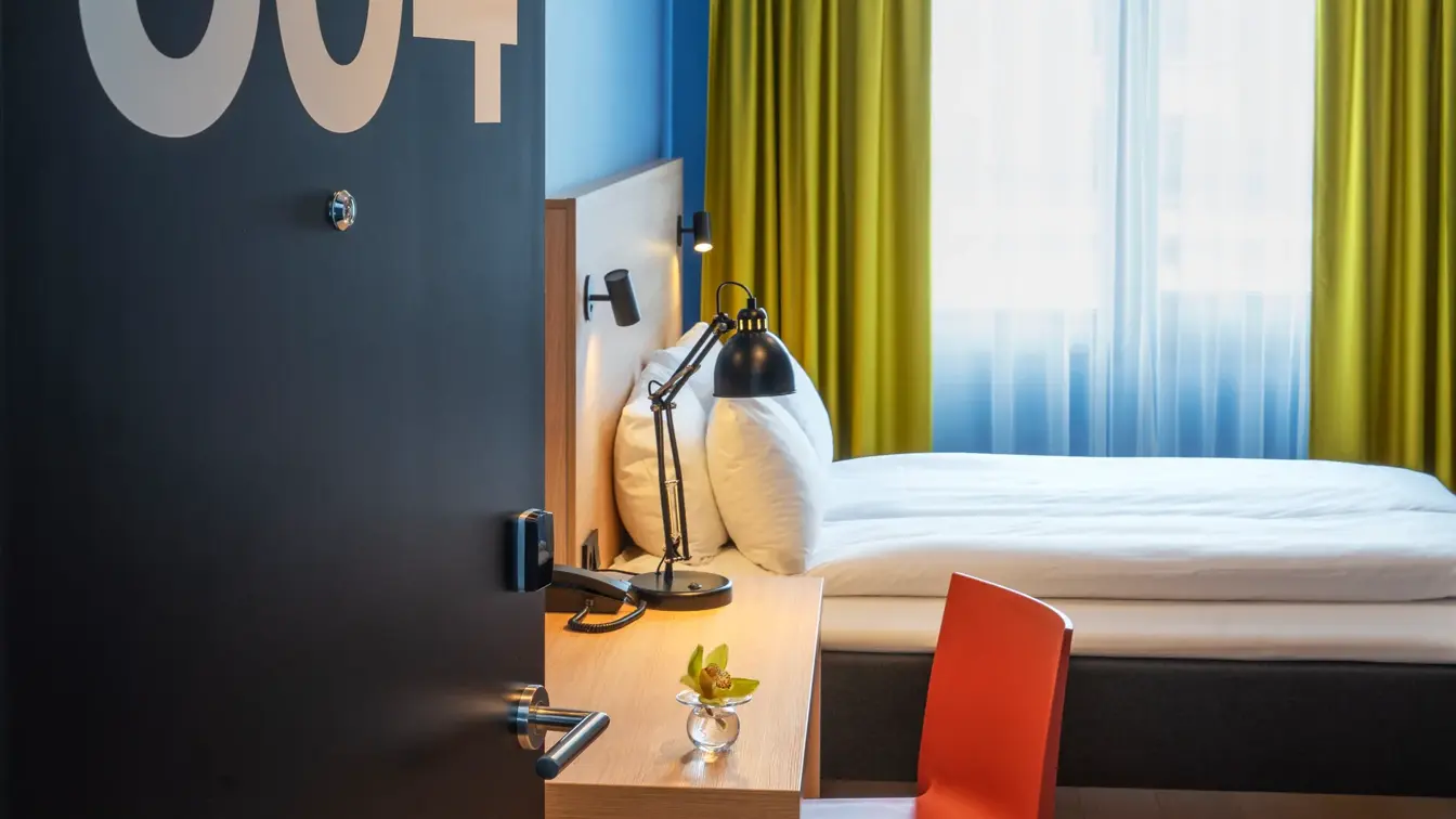 Åpen sort dør inn til rommet med romnummer 504 i stor hvit skrift. Skrivebord med rød stol, blå vegger, dobbeltseng og gule gardiner i dobbeltrom på Thon Hotel Astoria