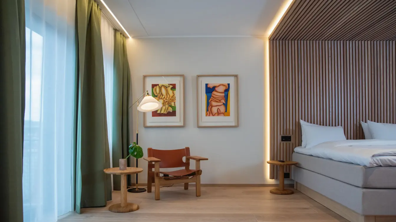 Store vinduer med grønne gardiner. Dobbeltseng med stripete tapet som sengegavl og i tak over sengen. To fargerike bilder på hvit vegg. En stol i træ og skinn og lite rundt bord.  I suite på Thon Hotel Norge i Kristiansand.
