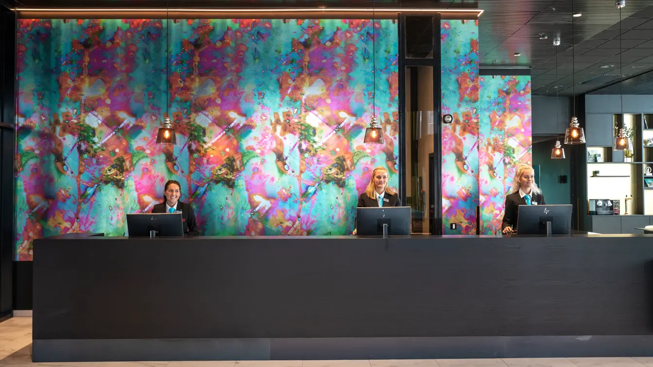 Resepsjonister bak resepsjonsdisk med fargerik tapet på Thon Hotel Storo