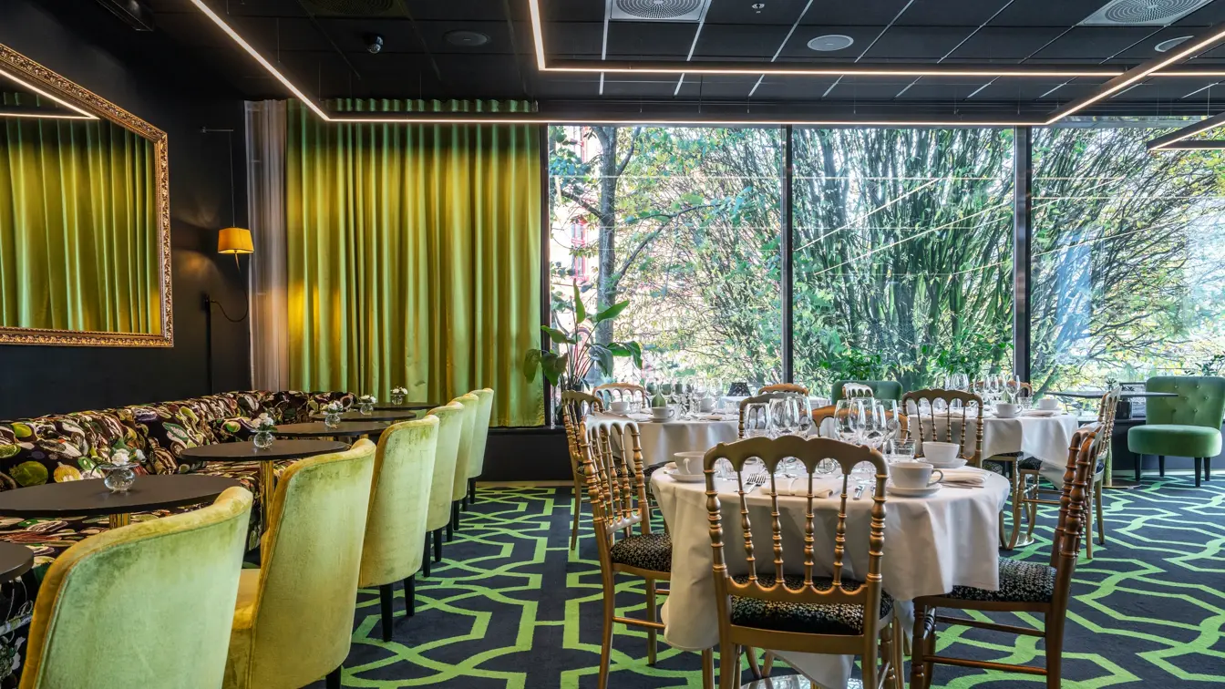 Lounge med grønt interiør, mønstrete sofa, grønne lenestoler, grønne gardiner, mønstret teppe, runde småbord dekket med hvit duk og servise.