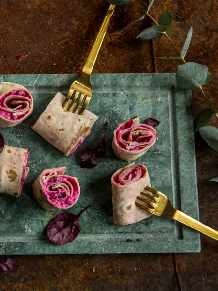 Lefseruller med rødbeter og lammerull sett ovenfra på et grønt marmorfat og gullgafler dandert rundt. 