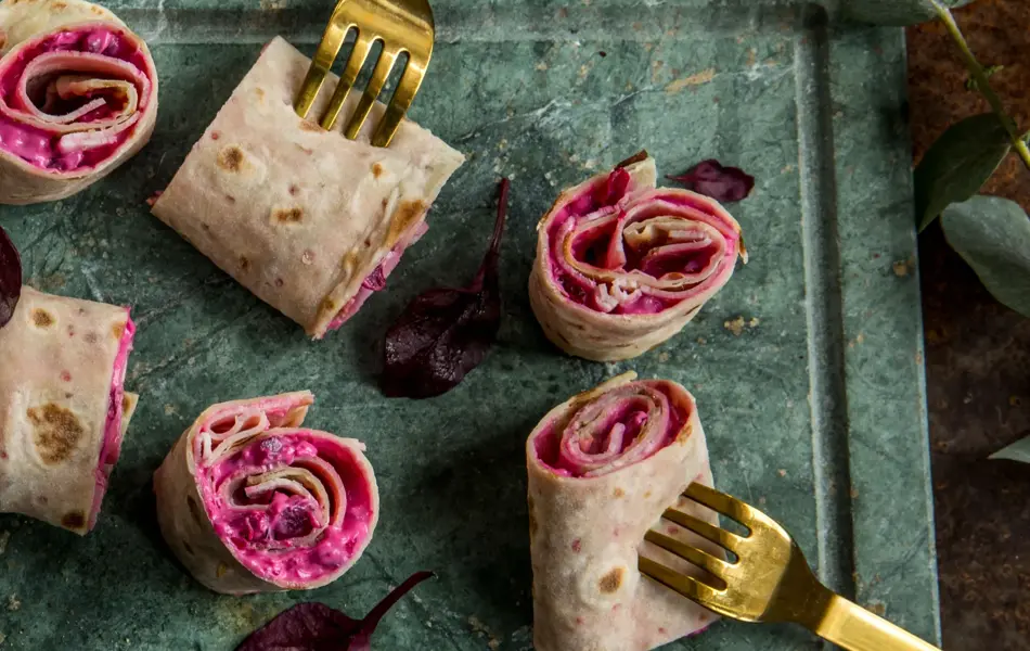 Lefseruller med rødbeter og lammerull sett ovenfra på et grønt marmorfat og gullgafler dandert rundt. 