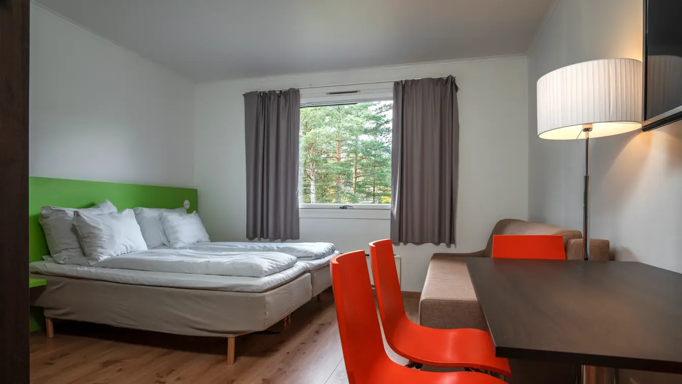 Studioleilighet med twin senger, sofa, vindu med grå gardiner, spisebord med rød stoler