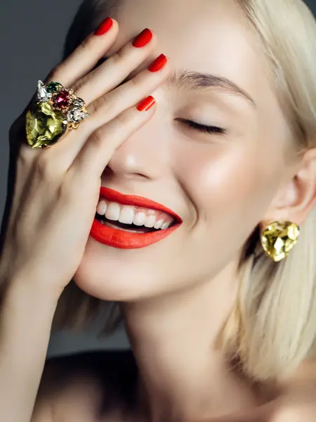 Nærbilde av smilende dame med rød lebestift og neglelakk, øredobb og ring i gull, holder den ene hånden opp foran ansiktet