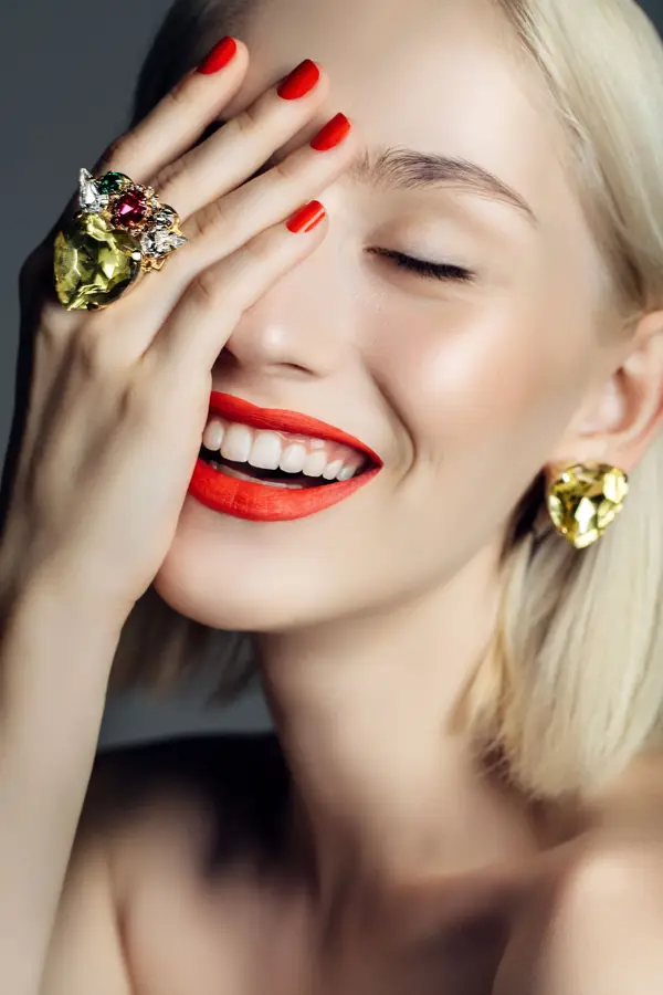 Nærbilde av smilende dame med rød lebestift og neglelakk, øredobb og ring i gull, holder den ene hånden opp foran ansiktet