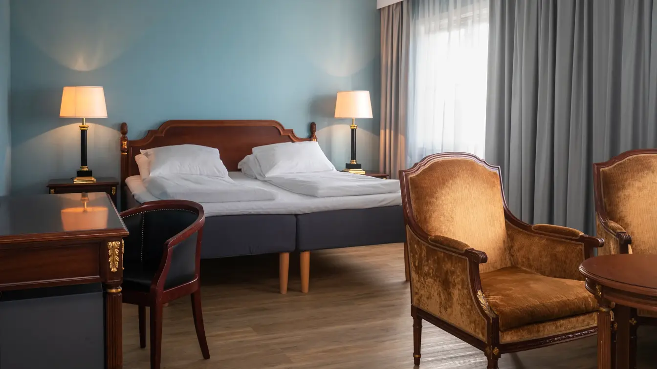 Dobbeltseng, parkettgulv, turkise vegger, vindu fra gulv til tak, grå gardiner, to sittestoler, skrivebord, stol. 