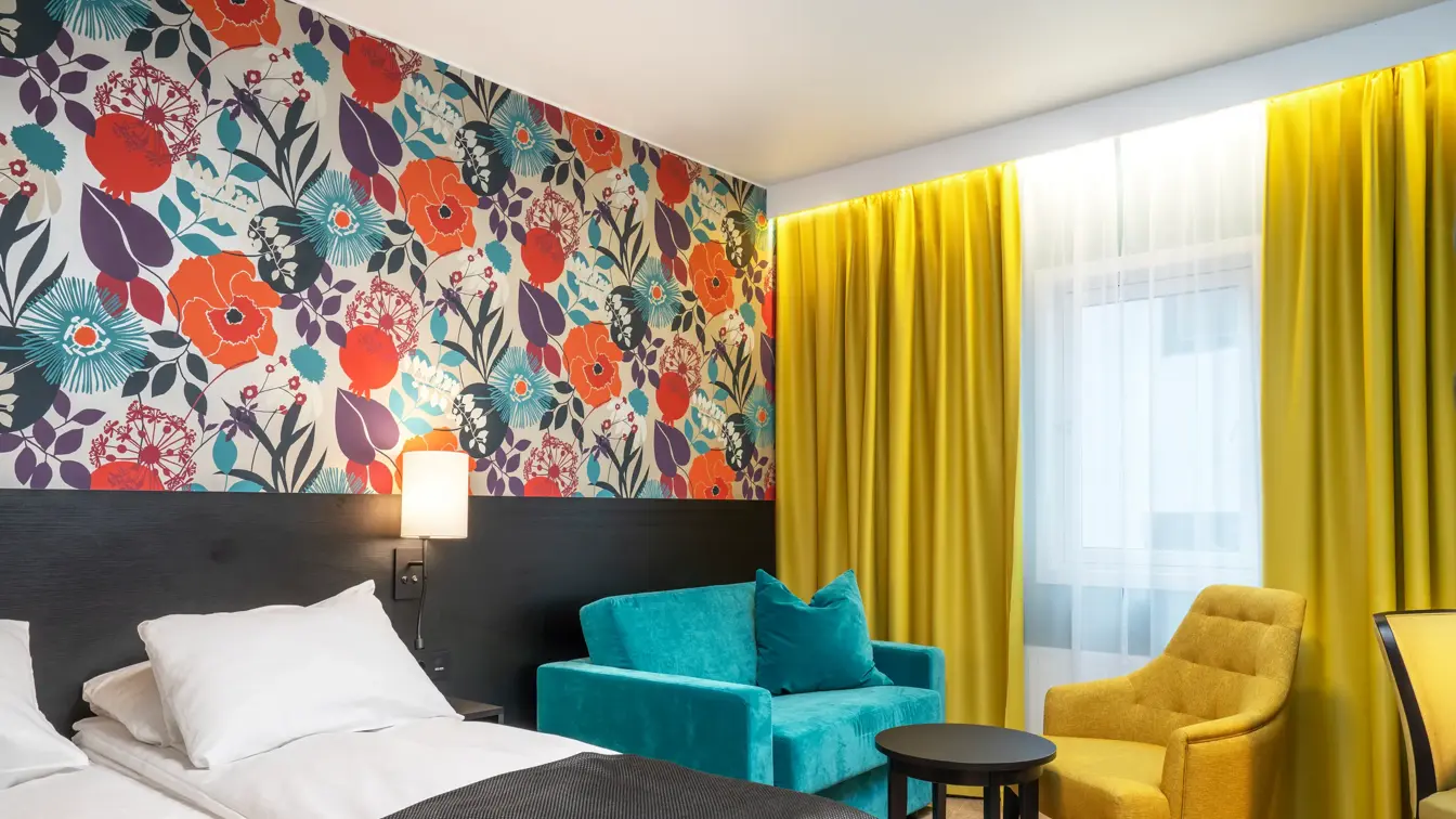 Blomstrende veggtapet, stort vindu med gule gardiner, blå og gul stol, og en dobbeltseng. 