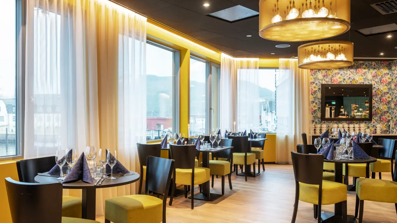 Svarte spisebord med svarte og gule stoler, Fem store vinduer, to store gule taklamper og en blomstrende tapetvegg