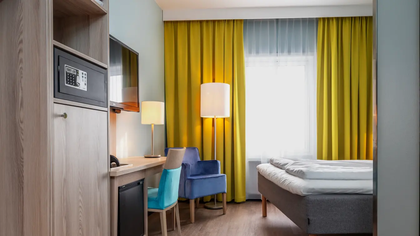 Dobbeltrom med dobbeltseng, skrivebord med turkis stol, lilla lenestol, gule gardiner, vegg hengt TV og safe i skap.
