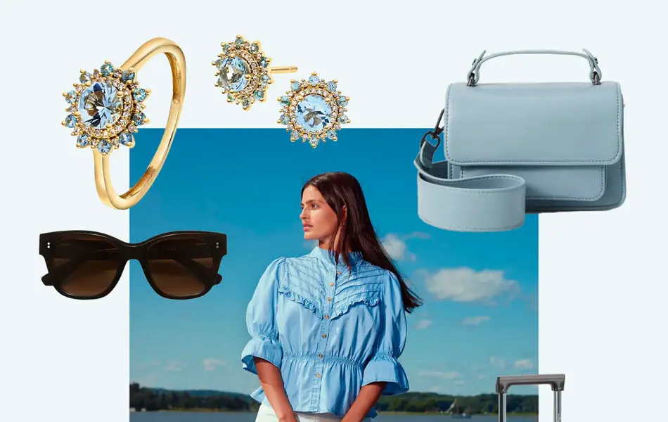 Gullring med stor blå sten, øredobber med blå sten, blå skulderveske, koffer, hvite sneakers, solbriller. Dame står på en brygge med blå bluse, hvite jeans, hvite pumps og liten blå veske.