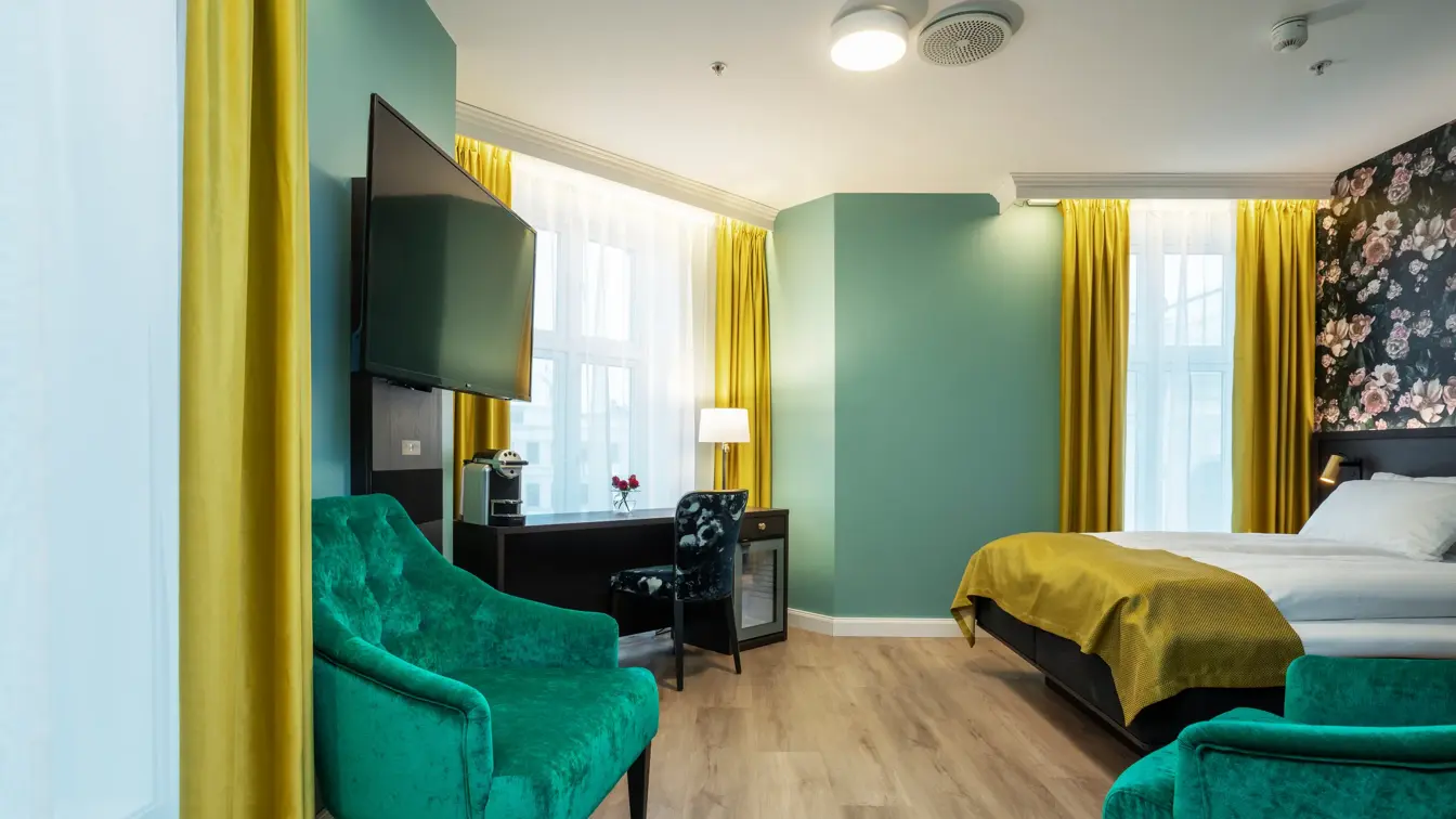 Store vinduer med gule gardiner fra tak til gulv. Veggehengt TV, to grønne lenestoler. Dobbeltseng med gult sengeteppe og mørkt skrivebord med mønstrete stol. I superior rom på Thon Hotel Høyers i Skien.