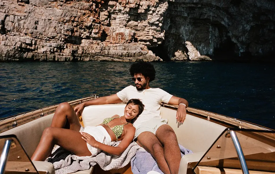 Dame med hvitt skjørt og grønn bikini hviler i fanget til mann i hvit t-skjorte og hvit shorts i en båt    + 