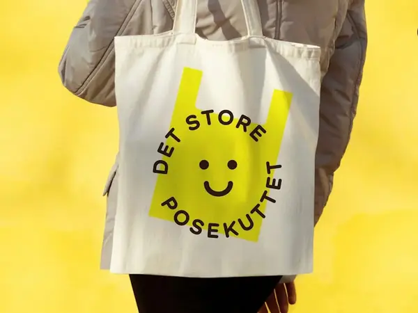 En person i beige jakke bærer på et lyst nett med en tegnet gul pose på. gul bakgrunn på bildet