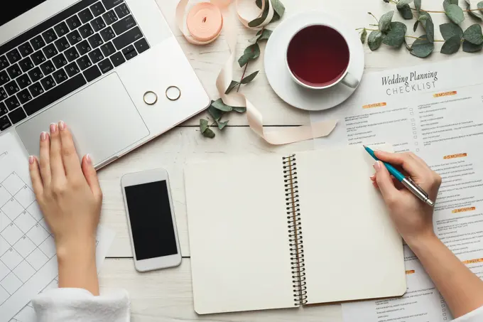 Wedding arrangement background. Female hands preparing for marriage, using laptop, smartphone, paper calendars and planners, top view. Bridal wallpaper with copy space on to do list