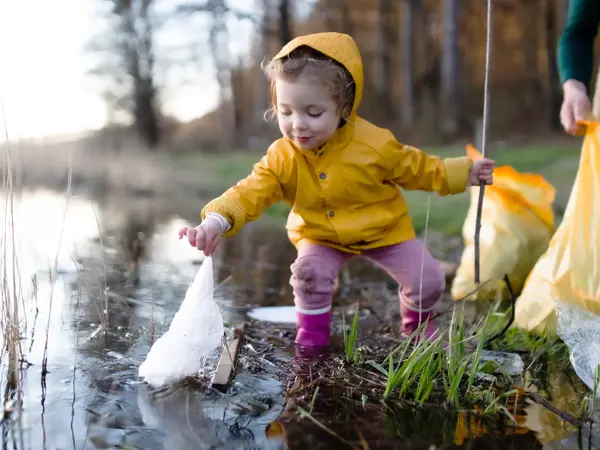Et barn i gul regnjakke som plukker en plastpose ut av et vann.