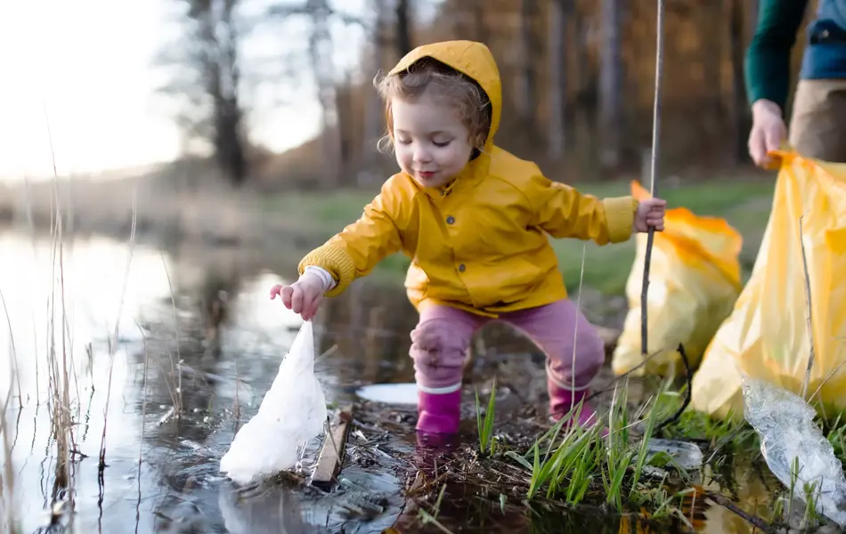 Et barn i gul regnjakke som plukker en plastpose ut av et vann.