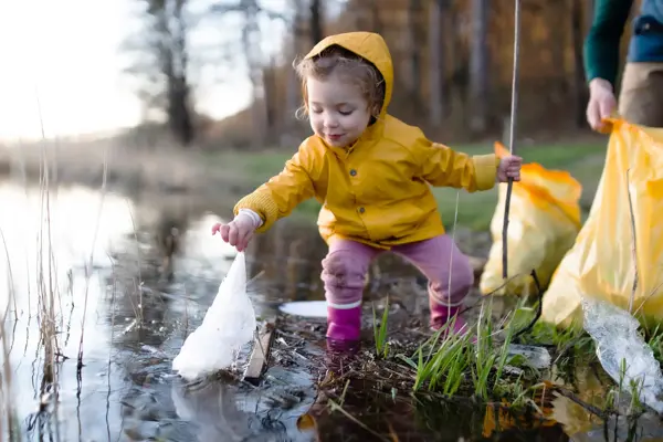 Et barn i gul regnjakke som plukker en plastpose ut av et vann.