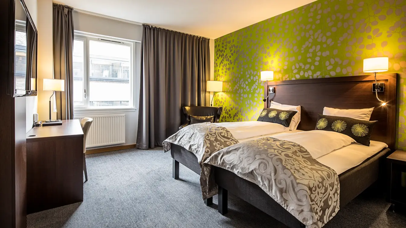 Grønn tapetvegg, dobeltseng, skrivebord, vindu, grå gardiner, to svarte stoler, 