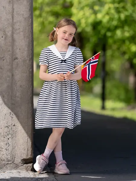 Jente står  ute i blå og hvitstripete matrosdress og rosa sneakers, holder et flagg.