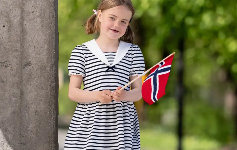 Jente står  ute i blå og hvitstripete matrosdress og rosa sneakers, holder et flagg.