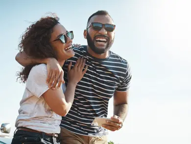 Mann med stripete t-skjorte og solbriller holder rundt dame med hvit t-skjorte og solbriller stående foran en bil