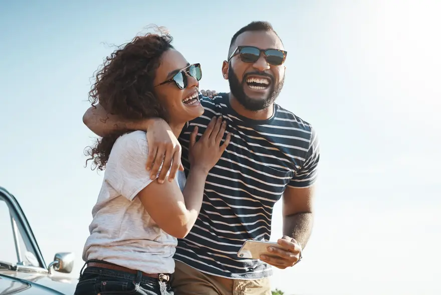 Mann med stripete t-skjorte og solbriller holder rundt dame med hvit t-skjorte og solbriller stående foran en bil