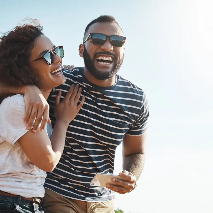 Mann med stripete t-skjorte og solbriller holder rundt dame med hvit t-skjorte og solbriller stående foran en bil