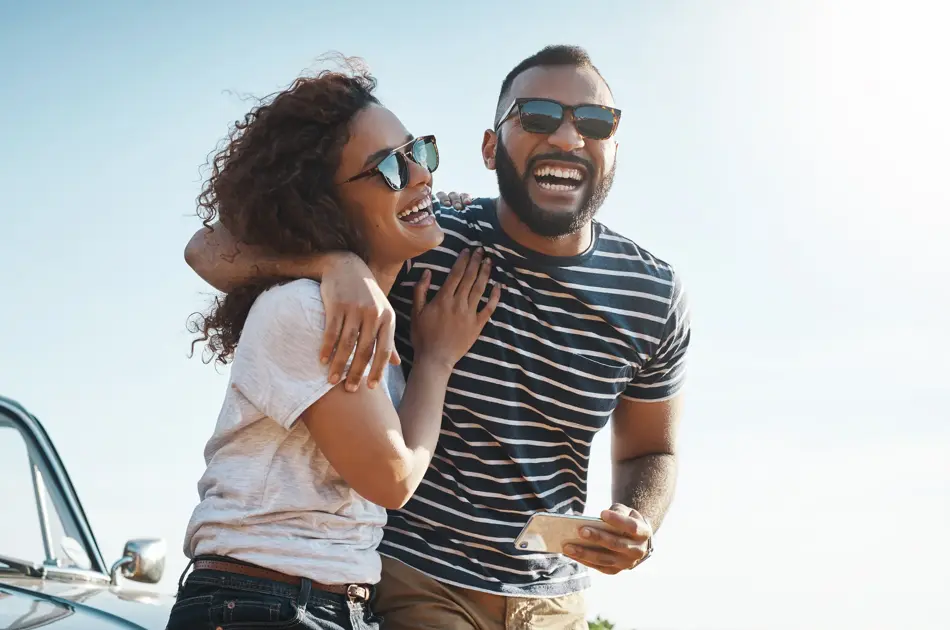 Mann med stripete t-skjorte og solbriller holder rundt dame med hvit t-skjorte og solbriller stående foran en bil
