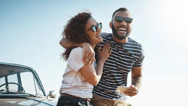 Mann med stripete t-skjorte og solbriller holder rundt dame med hvit t-skjorte og solbriller stående foran en bil