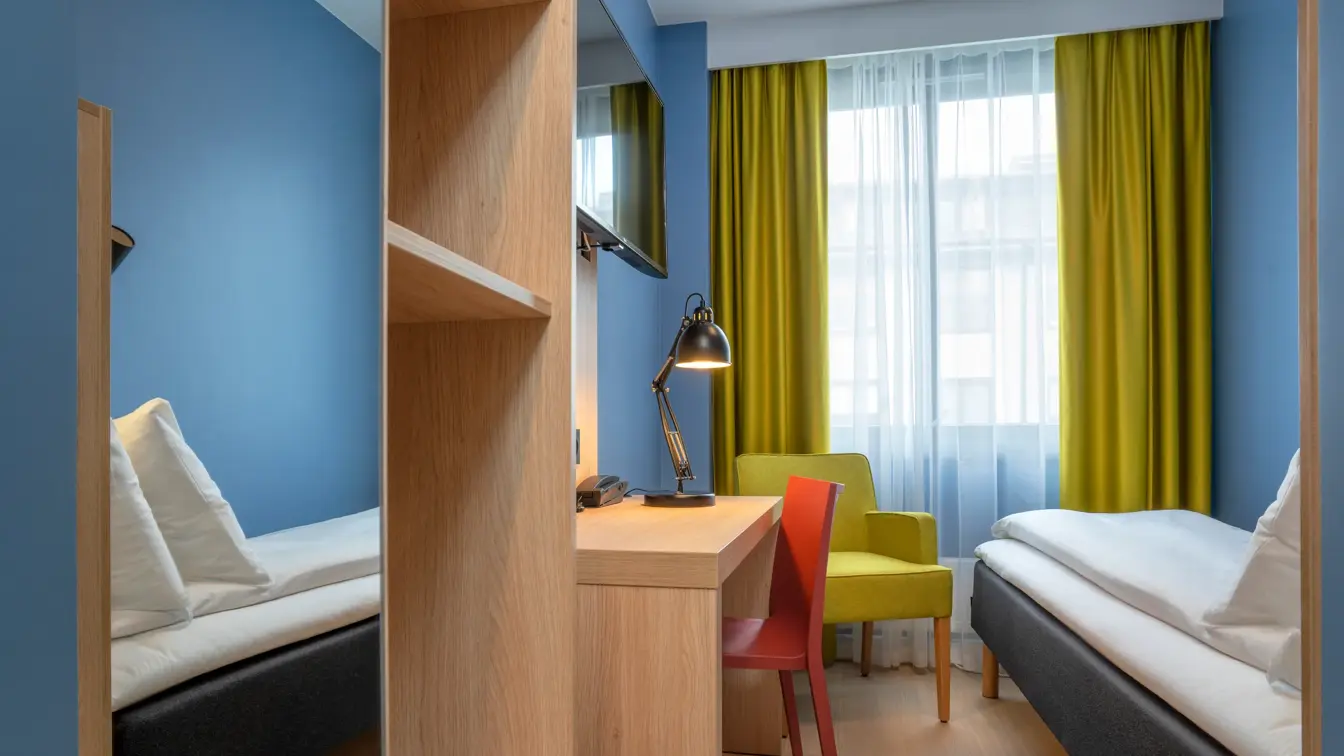 Speil, garderobe, skrivebord, TV, blå vegger, gul stol, gule gardiner og enkeltseng på lite enkeltrom på Thon Hotel Astoria