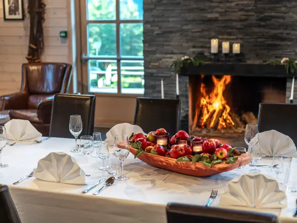 Oppdekket bord foran peis med flammer, hvit duk, brettede servietter, glass, bestill og et stort fat med epler i midten.
