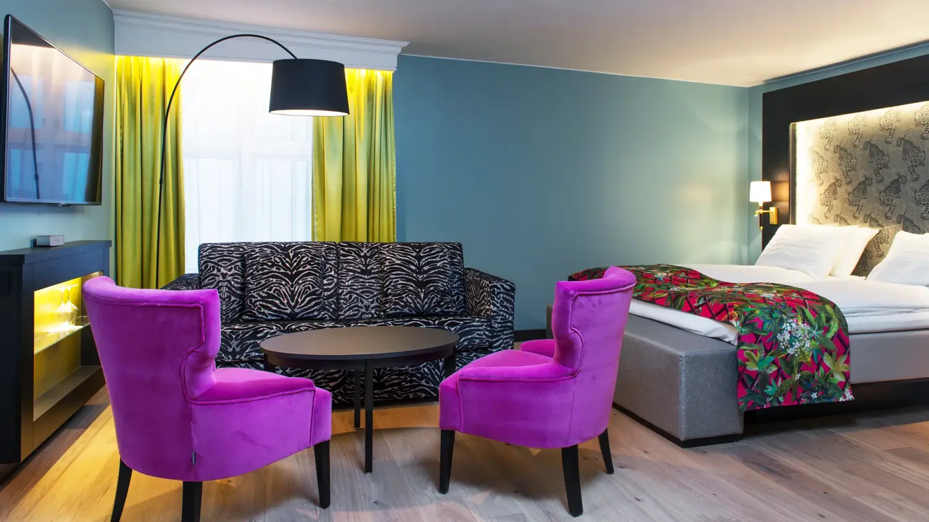 Dobbeltseng med mønstrede sengeteppe og lilla stol, zebramønstret sofa og to lilla stoler i sittegruppe, TV og gule gardiner på buisness room på Thon hotel Terminus i Oslo sentrum nær Jernbanetorget