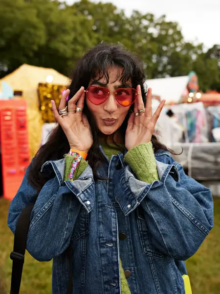 Dame med rosa hjertesolbriller og blå jeansjakke poserer foran kamera på festival  