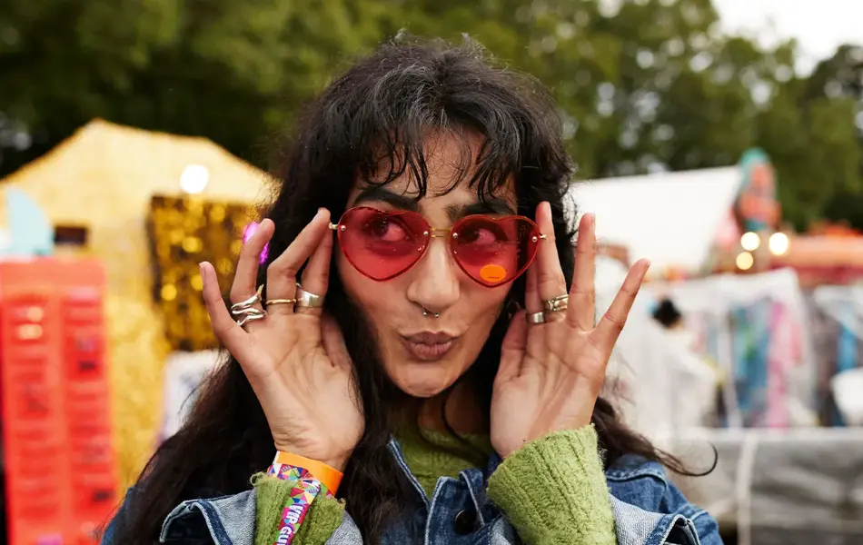 Dame med rosa hjertesolbriller og blå jeansjakke poserer foran kamera på festival  