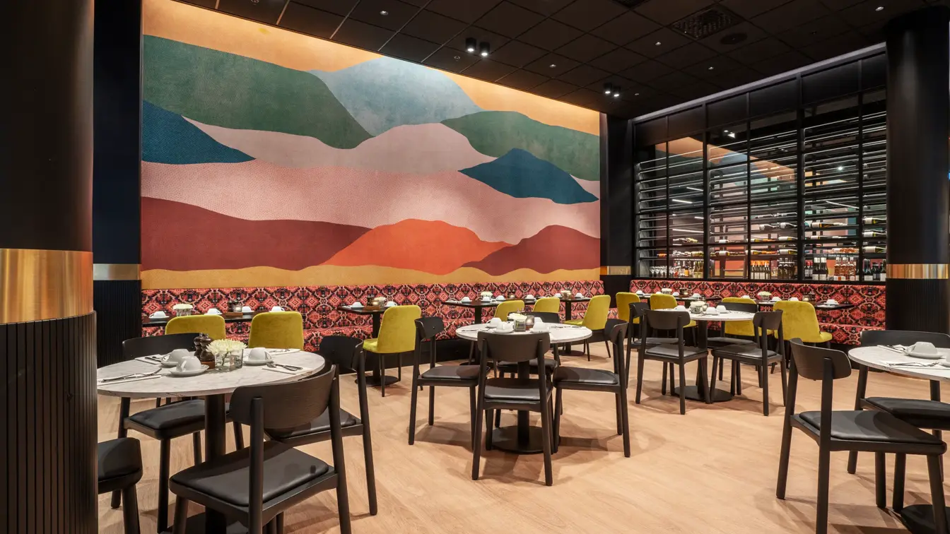 Fargerik vegg i bakgrunnen, rød sofa med sort og hvit mønster, kvadratiske og runde spisebord, gule og brune stoler.