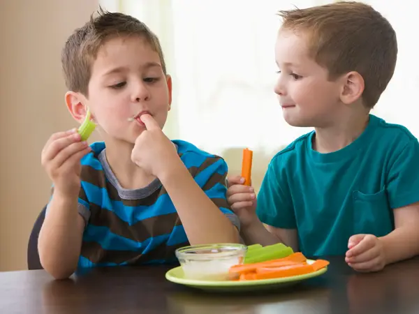 To gutter spiser grønnsaker og dipp ved et bord.