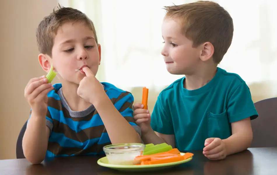 To gutter spiser grønnsaker og dipp ved et bord.