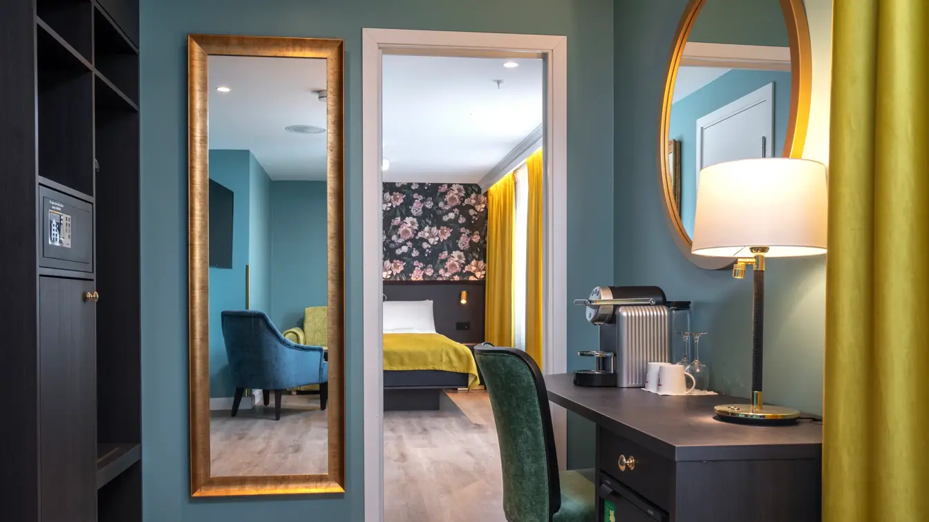 Skrivebord, grønn stol, kaffemaskin, rundt veggspeil, parkettgulv, vindu, gule gardiner, dobbeltseng