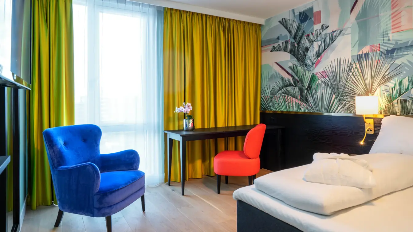 Del av dobbeltseng, arbeidspult og stor blå lenestol, store lyse vinduer og gule gardiner på dobbeltrom på Thon Hotel Storo i Oslo