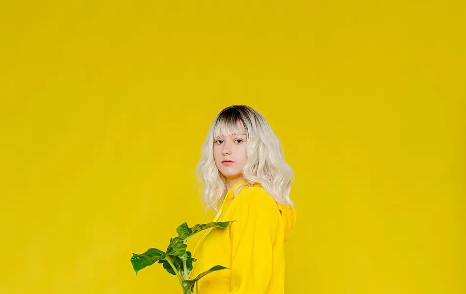 Jente med hvite sko, knallgule bukser og knallgul genser, står i helt gult rom og ser mot kamera mens hun holder en grønn potteplante i hendene.