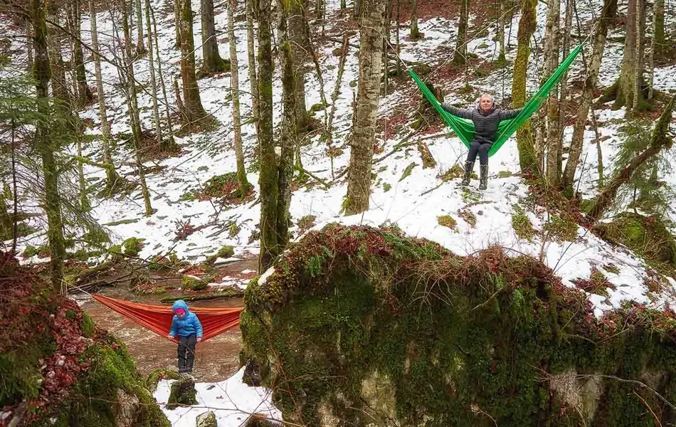 To personer i hver sin hengekøye mellom trær i delvis snødekket skog